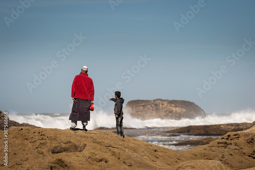 Father and son on rocks near to ocean with big waves and blue sky in background and seagulls flying around. Essaouira, Morocco