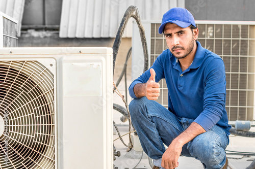 a professional electrician man is fixing a heavy duty unit of central air conditioning system by his tools on the roof top and wearing blue color of uniform and white cap
