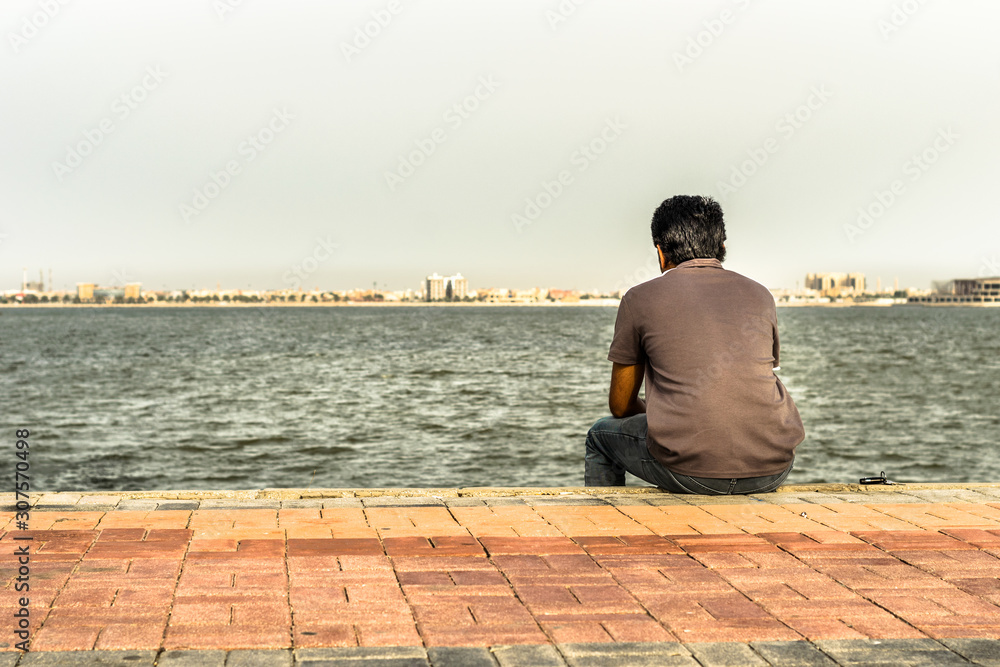 a lonely man is sitting on the rocks after divorce at seaside wearing grey t shirt and blue jeans in the bright cloudy day