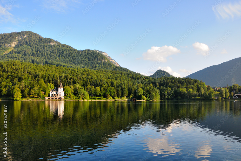 Fototapeta premium A large white house in the form of a castle on the shore of the lake. Green mountains in the background. Blue sky, summer day.
