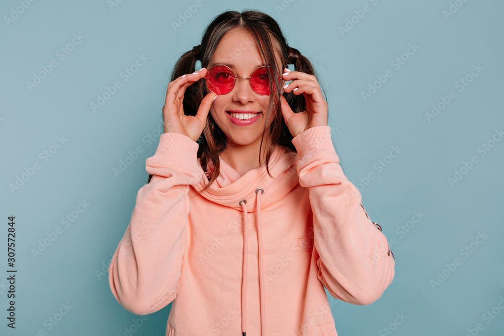 Close up portrait of smiling happy girl in round pink glasses and wearing pink blouse looking at camera over blue background Close up portrait of smiling happy girl in round pink glasses and wearing pink blouse looking at camera over blue background
