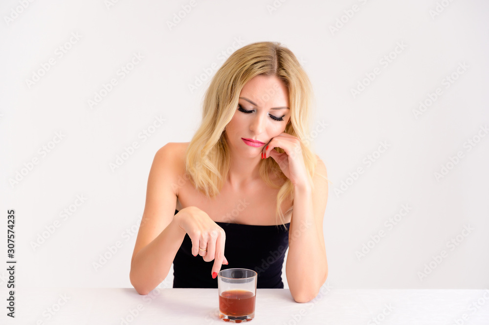 Portrait of a pretty blonde model woman with excellent make-up, beautiful hair and clean skin on a white background sitting at a table with a glass of whiskey in her hands. Portrait of a pretty blonde model woman with excellent make-up, beautiful hair and clean skin on a white background sitting at a table with a glass of whiskey in her hands.