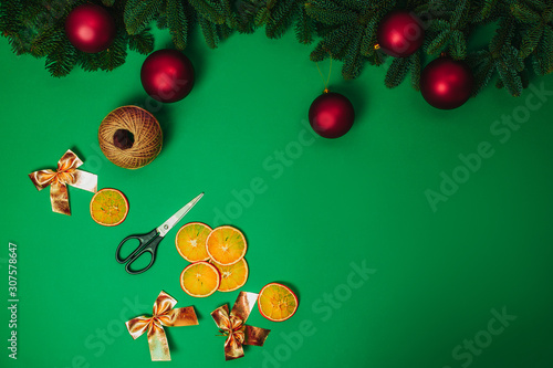 Christmas hand made how to make Christmas wreath. Scissors, rope, red christmas balls, golden bows with fir branches and dried oranges on the green background.