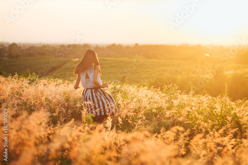Trendy girl in stylish summer dress feeling free in the field with flowers in sunshine.