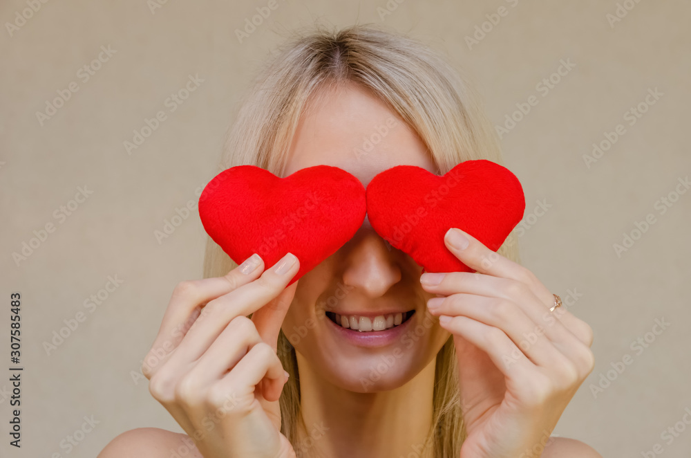 Lifestyle concept: attractive blonde woman with a radiant smile, having two small red hearts in her hands, closing her eyes with heart symbols, standing on a light background. Valentine's day Lifestyle concept: attractive blonde woman with a radiant smile, having two small red hearts in her hands, closing her eyes with heart symbols, standing on a light background. Valentine's day