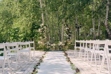 Place for wedding ceremony in garden, copy space. Wedding arch decorated with flowers and white chairs on each side of archway outdoors. Wedding setting