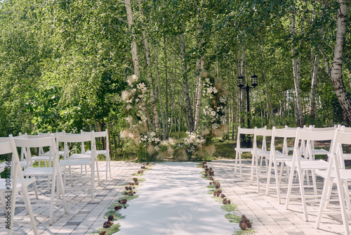 Place for wedding ceremony in garden, copy space. Wedding arch decorated with flowers and white chairs on each side of archway outdoors. Wedding setting
