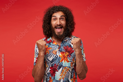 Overjoyed attractive young brunette bearded man in flowered shirt raising fists happily and looking cheerfully to camera with wide eyes and mouth opened, isolated over red background