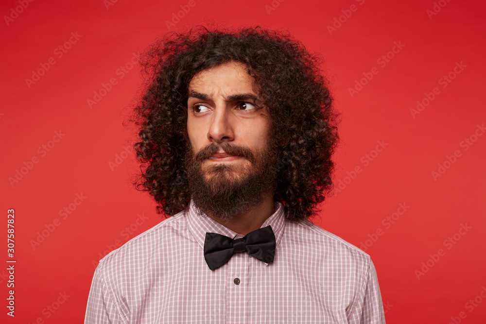 Portrait of young curly dark haired bearded male raising eyebrows and keeping lips folded while looking seriously aside, dressed in formal clothes while standing against red background Portrait of young curly dark haired bearded male raising eyebrows and keeping lips folded while looking seriously aside, dressed in formal clothes while standing against red background