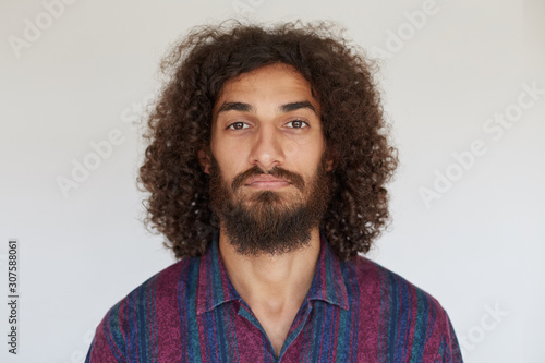 Portrait of young handsome brown-eyed bearded male with dark curly hair looking at camera with calm face, keeping lips folded while standing against white background