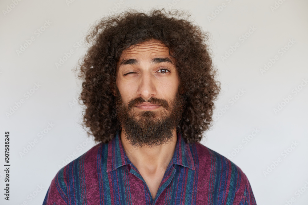Portrait of pretty young dark haired curly bearded man with folded lips keeping eye closed while looking to camera, wearing casual clothes while standing over white background