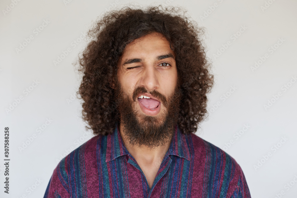 Indoor shot of young brunette curly male with lush beard giving wink to camera with wide mouth opened, wearing striped multi-colored shirt against white background Indoor shot of young brunette curly male with lush beard giving wink to camera with wide mouth opened, wearing striped multi-colored shirt against white background