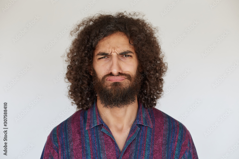 Fototapeta premium Displeased young pretty bearded male with curly dark hair looking to camera with pout, frowning eyebrows with folded lips against white background, wearing striped casual shirt