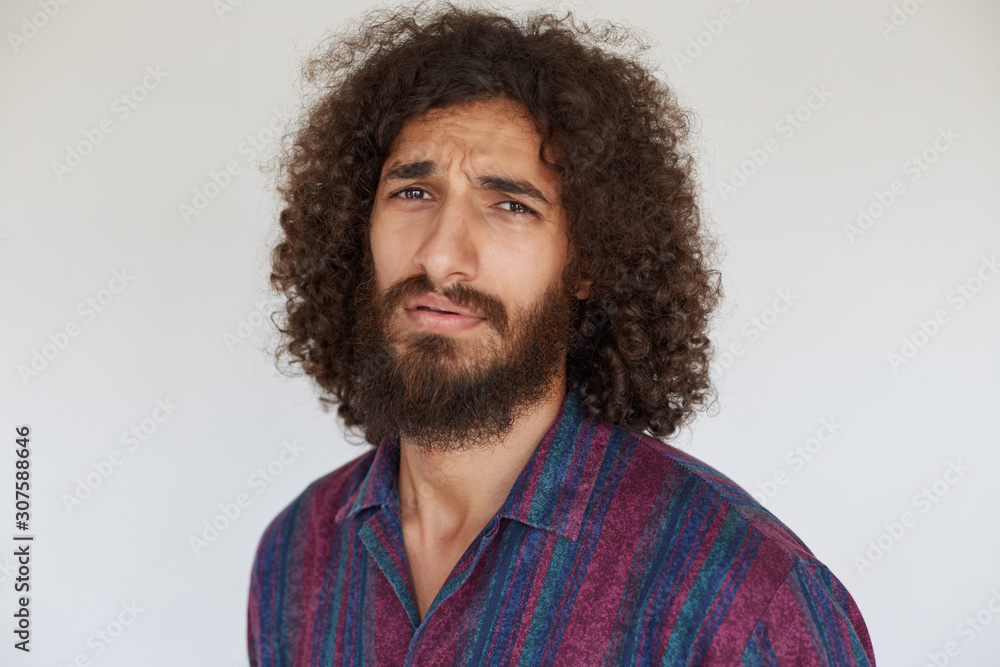 Discontented young dark haired curly male with lush beard frowning eyebrows with pout while standing against white background in striped multi-colored shirt Discontented young dark haired curly male with lush beard frowning eyebrows with pout while standing against white background in striped multi-colored shirt