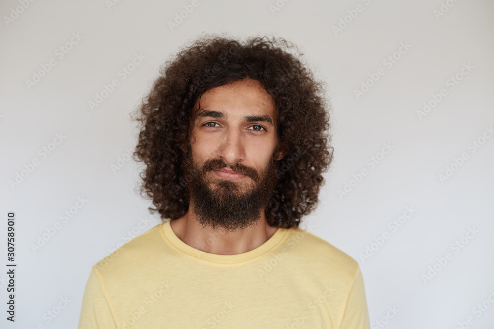 Indoor photo of pleasant looking positive young bearded man with brown curly hair smiling slightly while posing over white background, wearing casual clothes Indoor photo of pleasant looking positive young bearded man with brown curly hair smiling slightly while posing over white background, wearing casual clothes