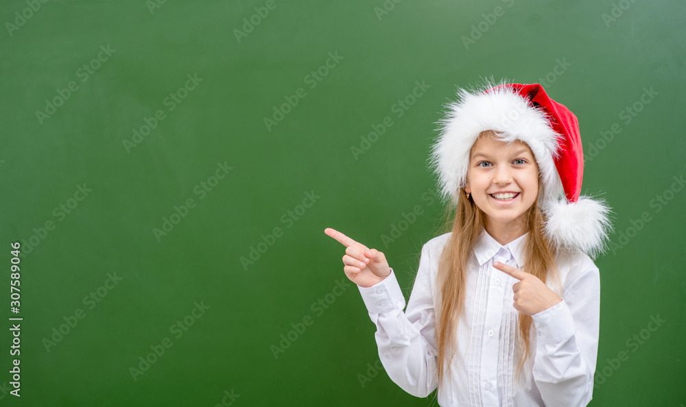 Smiling girl wearing a red christmas hat points away on empty chalkboard. Space for text Smiling girl wearing a red christmas hat points away on empty chalkboard. Space for text
