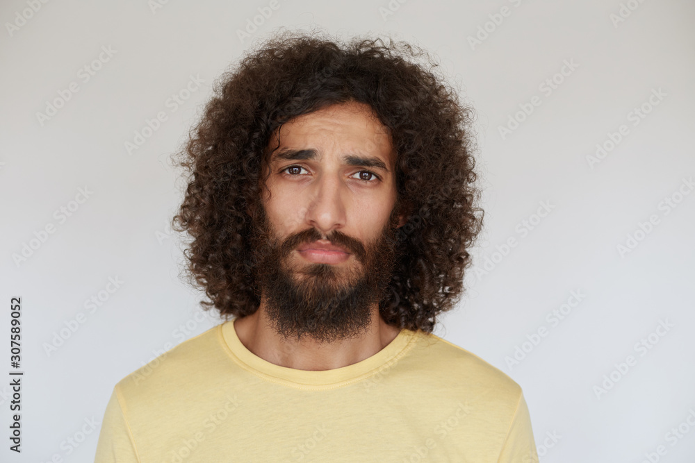 Upset beautiful brown-eyed young bearded man with dark curly hair looking sadly to camera and keeping his lips folded, standing against white background in casual clothes Upset beautiful brown-eyed young bearded man with dark curly hair looking sadly to camera and keeping his lips folded, standing against white background in casual clothes
