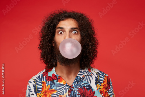 Astonished attractive young brunette curly man with beard wearing multi-colored flowered shirt while posing over red background, rounding eyes and inflating bubble with pink gum photo