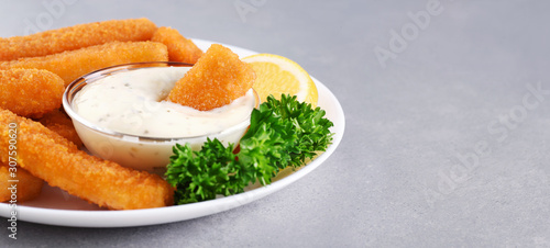 Fish fingers sticks on a plate with parsley, lemon and tartar sauce on a gray texture.