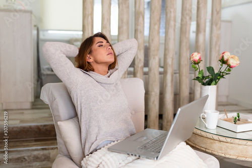 Overworked young woman sitting in her home with a laptop in front of her stretching her arms above her head with eyes closed to relax muscles, reduce tension, improve vision, photo