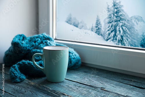 Winter background - cup with candy cane, woolen scarf and gloves on windowsill and winter scene outdoors. Still life with concept of spending winter time at cozy home with cold weather outdoors