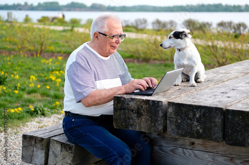 active old age, technology, senior people, lifestyle, distance learning. Happy Senior man 70-75 years old sitting in summer park with dog jack russell terrier and uses laptop computer.