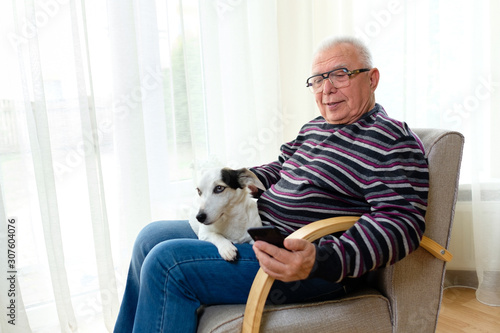 Senior man 70-75 years old sitting on armchair at home, reading, watching and relaxing with smartphone with his best friend, a dog Jack russell terrier.
