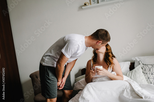 Loving husband carries coffee for his happy wife in the bed, in a big bedroom, side view. The Valentine's Day Concept. Celebrating holiday. Lifestyle. Women's Day, eighth of March. Young couple kiss.