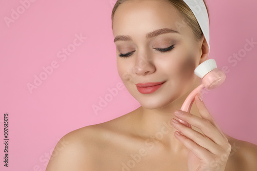 Young woman washing face with cleansing brush on pink background. Cosmetic product