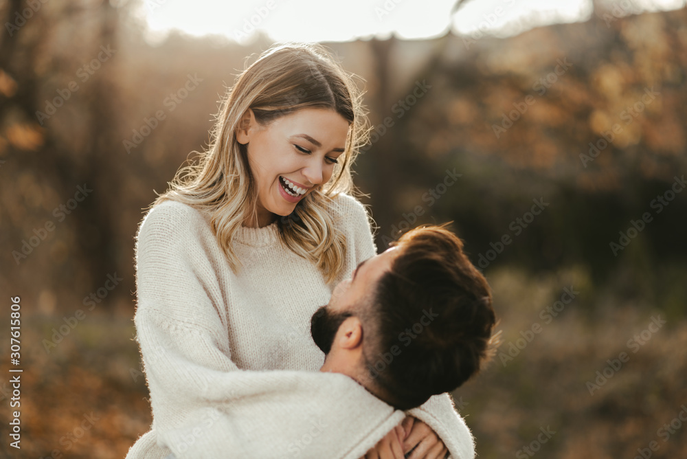 Young cheerful couple having fun in nature during autumn. Woman being carried by her boyfriend - obrazy, fototapety, plakaty 