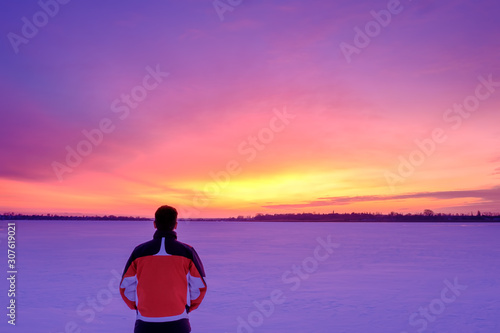 A man stands with his back to the camera on the snowy surface of a frozen lake and watches colorful twilight.