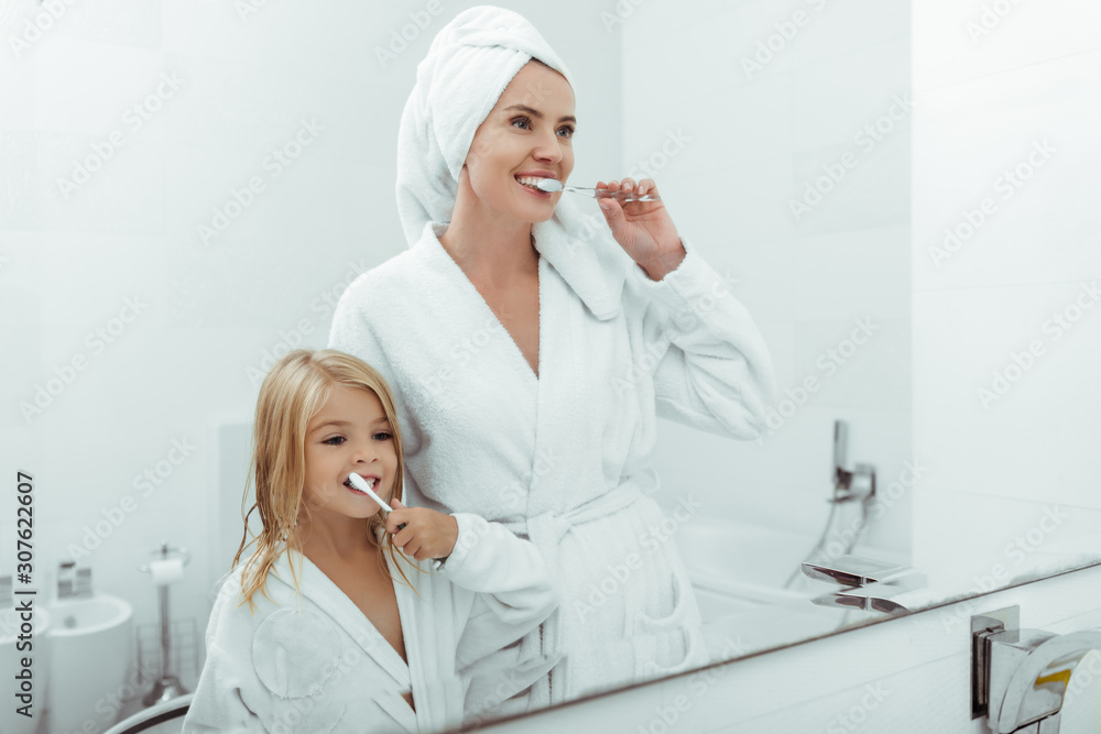 happy mother and daughter in bathrobes brushing teeth near mirror happy mother and daughter in bathrobes brushing teeth near mirror