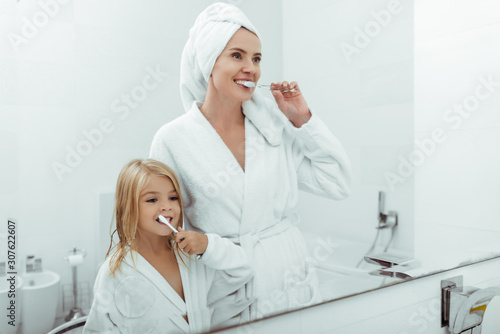 happy mother and daughter in bathrobes brushing teeth near mirror