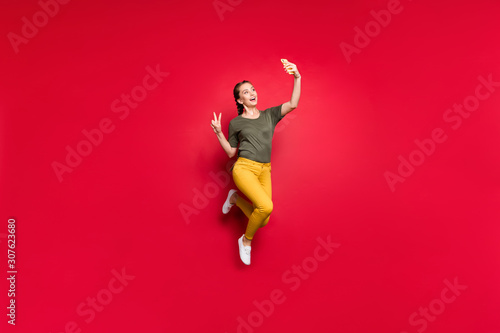 Full body photo of crazy funny lady jumping high making selfies showing v-sign symbol cheerful mood wear casual yellow pants green t-shirt isolated red color background