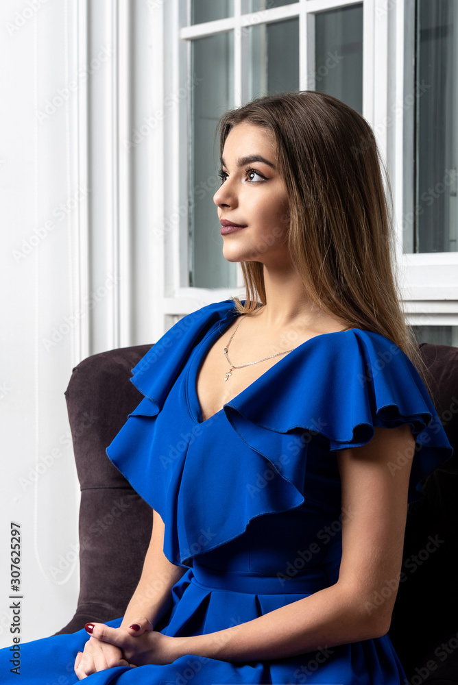 Portrait of beautiful young girl in blue dress sitting at home and looking at the light Portrait of beautiful young girl in blue dress sitting at home and looking at the light