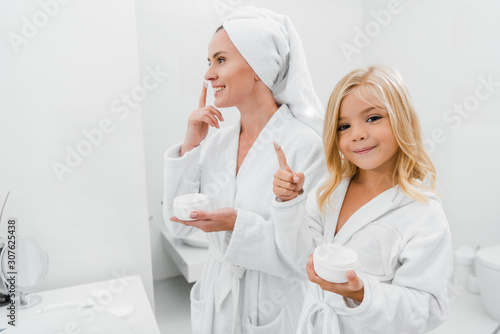 cheerful kid with face cream on finger near attractive mother in bathroom