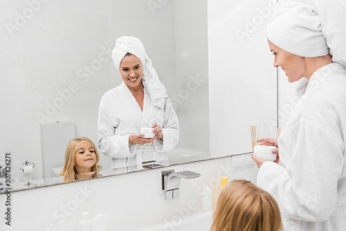 selective focus of happy mother holding container with cosmetic cream near cute daughter