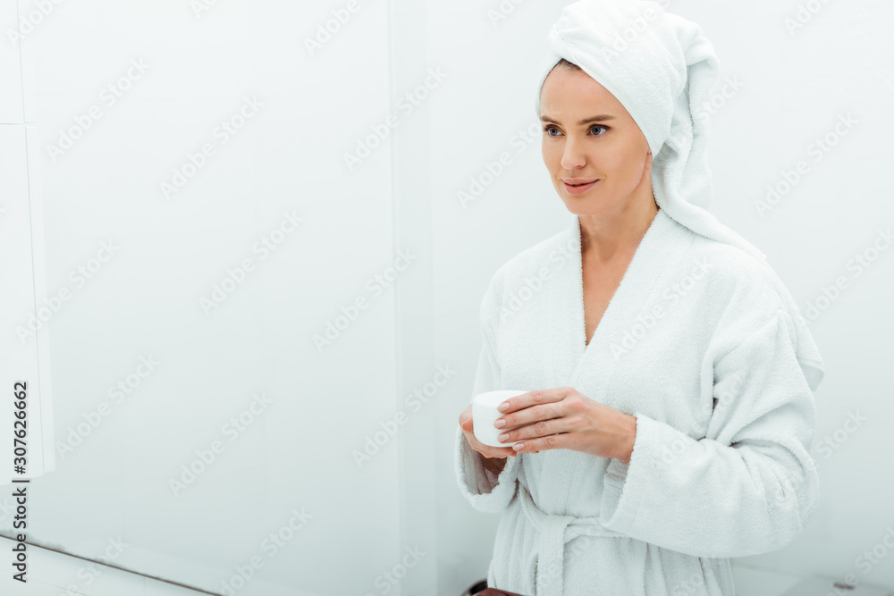 beautiful woman in bathrobe and towel holding container with face cream in bathroom beautiful woman in bathrobe and towel holding container with face cream in bathroom