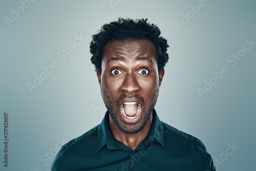 Surprised young African man looking at camera and keeping mouth open while standing against grey background