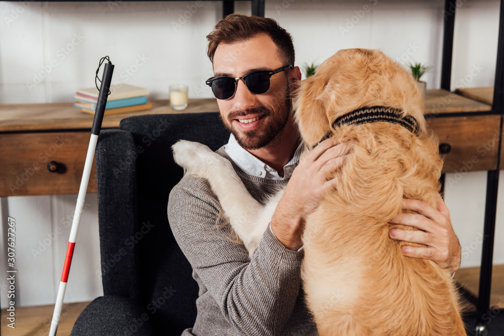 Smiling blind man sitting in armchair and hugging golden retriever at home - obrazy, fototapety, plakaty 