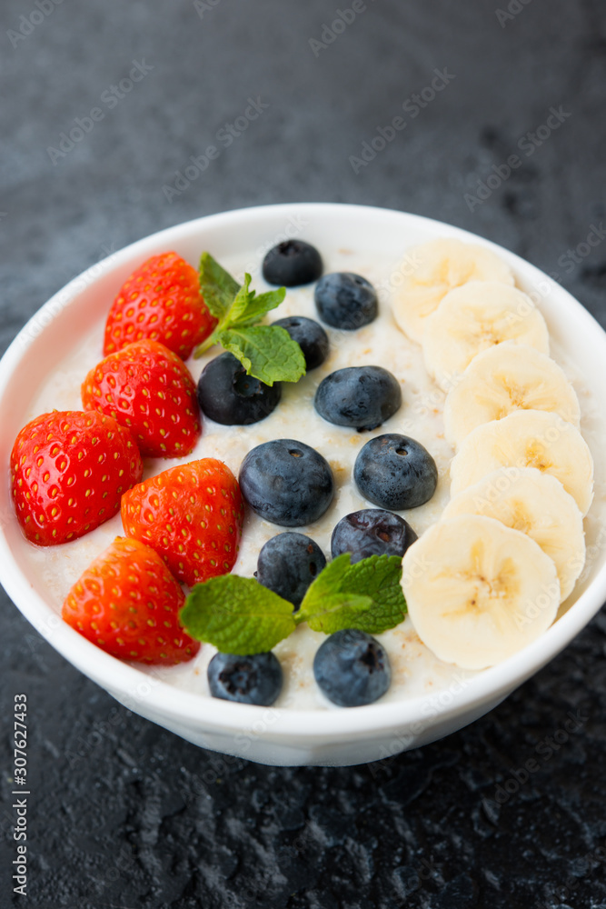 Delicious, healthy and balanced Breakfast of oatmeal with berries and fruit in a plate on the left on a concrete background. Vertical orientation Delicious, healthy and balanced Breakfast of oatmeal with berries and fruit in a plate on the left on a concrete background. Vertical orientation