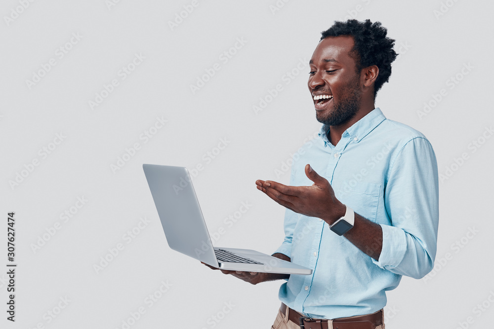 Handsome young African man using laptop and smiling while standing against grey background Handsome young African man using laptop and smiling while standing against grey background