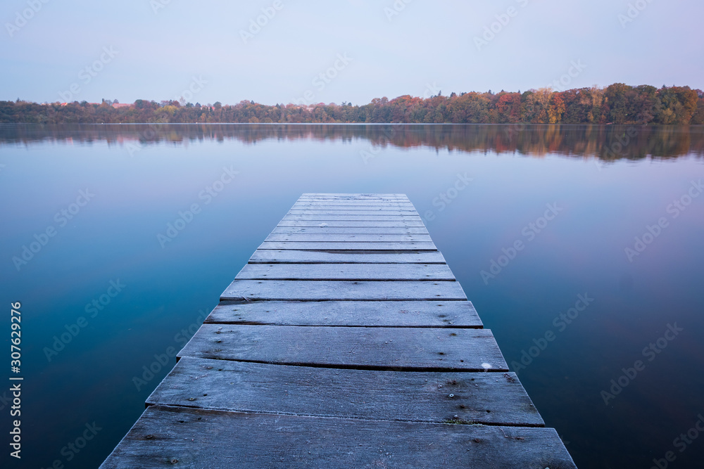 Obraz premium Old, wooden pier with frost surface into blue, calm lake on cloudy autumn morning with forest in background, Eutin, Schleswig-Holstein, Germanywinterday