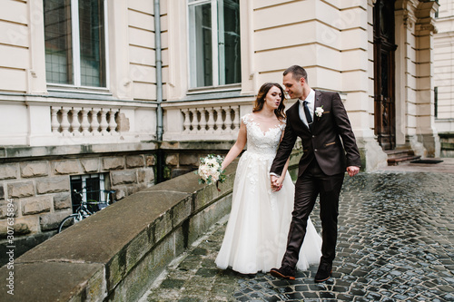 Portrait the groom in wedding suit and the bride in dress walking near ancient restored architecture, old building, old house outside, vintage palace outdoor. photo