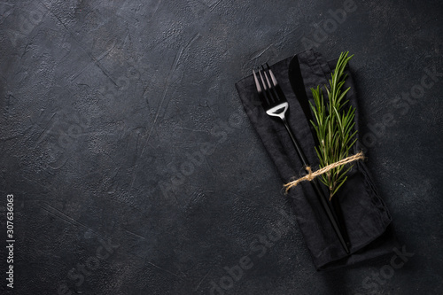 Black cutlery and napkin with a sprig of rosemary on black stone table top view.