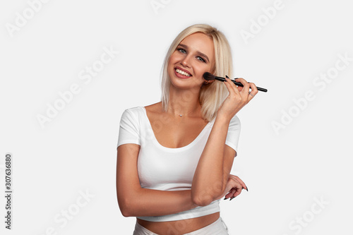 Beautiful blonde girl applies skin tone with Brush. Sexy woman portrait isolated on white background. Perfect Makeup. Skincare Foundation. Pretty girl with perfect skin smiling. Brushes Makeup Artist.