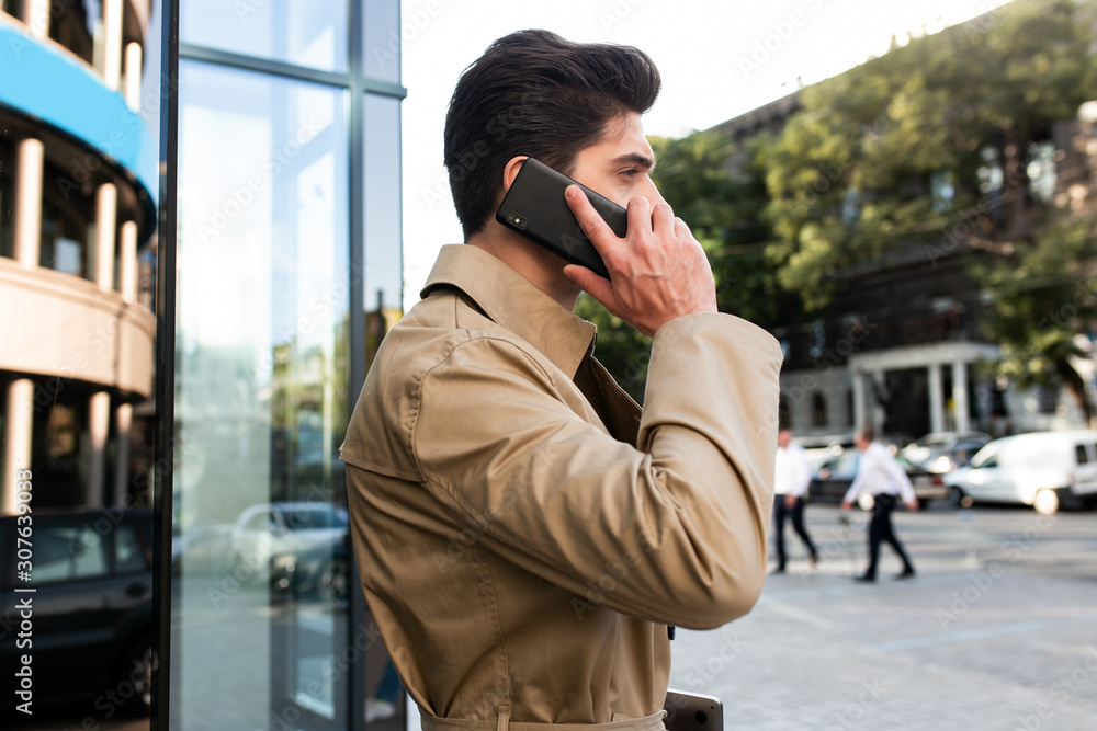 Side view of young stylish man in trench coat talking on cellphone on street Side view of young stylish man in trench coat talking on cellphone on street