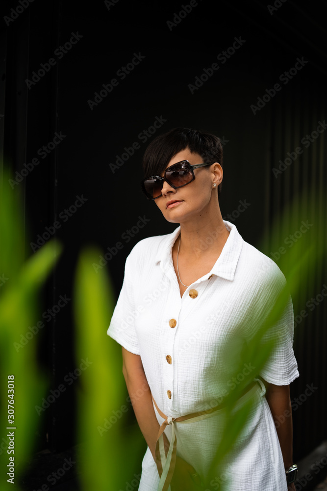Beautiful woman with short brunette hair in white clothes and sunglasses. Fashion street photography. Fashion model posing on dark background.