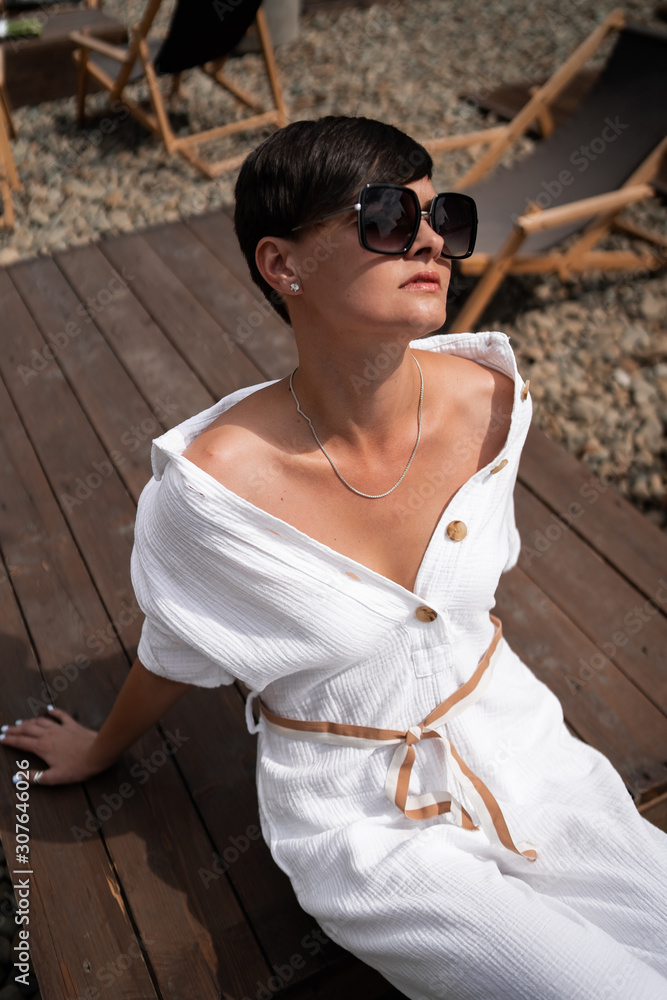 Beautiful woman with short brunette hair in white clothes and sunglasses. Fashion street photography. Fashion model is sitting and relax on brown wood background. sunglasses shop