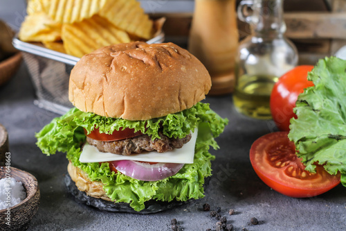Photo of fresh burger on wooden cutting board on dark background..Homemade hamburger with beef, onion, tomato, lettuce and cheese. Homemade fast food. Dark textured background. Copy space. Image.